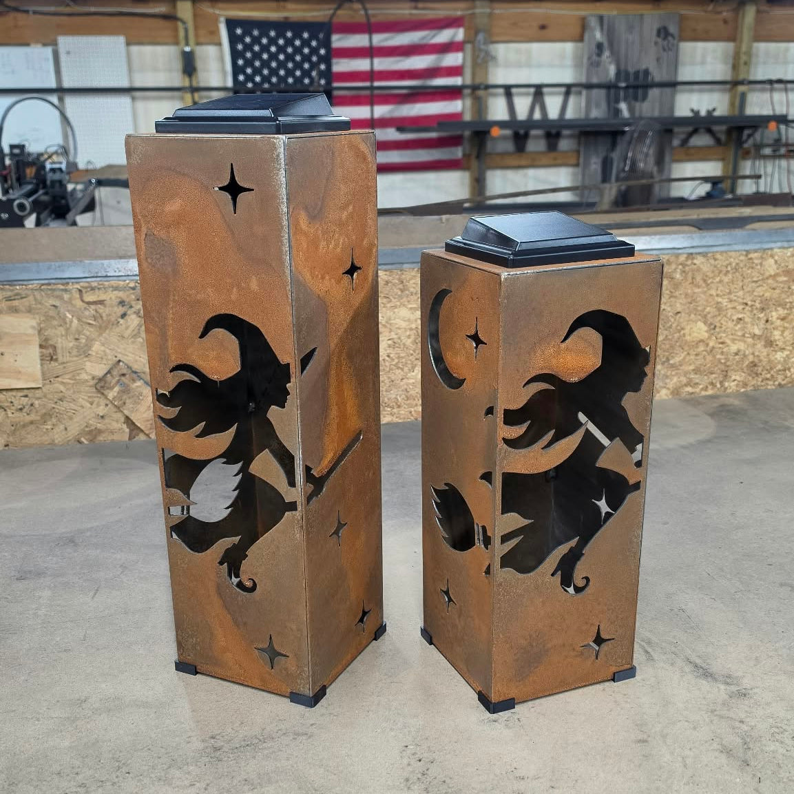 Two tall rectangular metal lanterns with a rustic copper-brown finish, featuring black cutout silhouettes of witches flying on broomsticks under a crescent moon and stars. The lanterns are photographed in a workshop setting with a concrete floor and wooden ceiling beams.