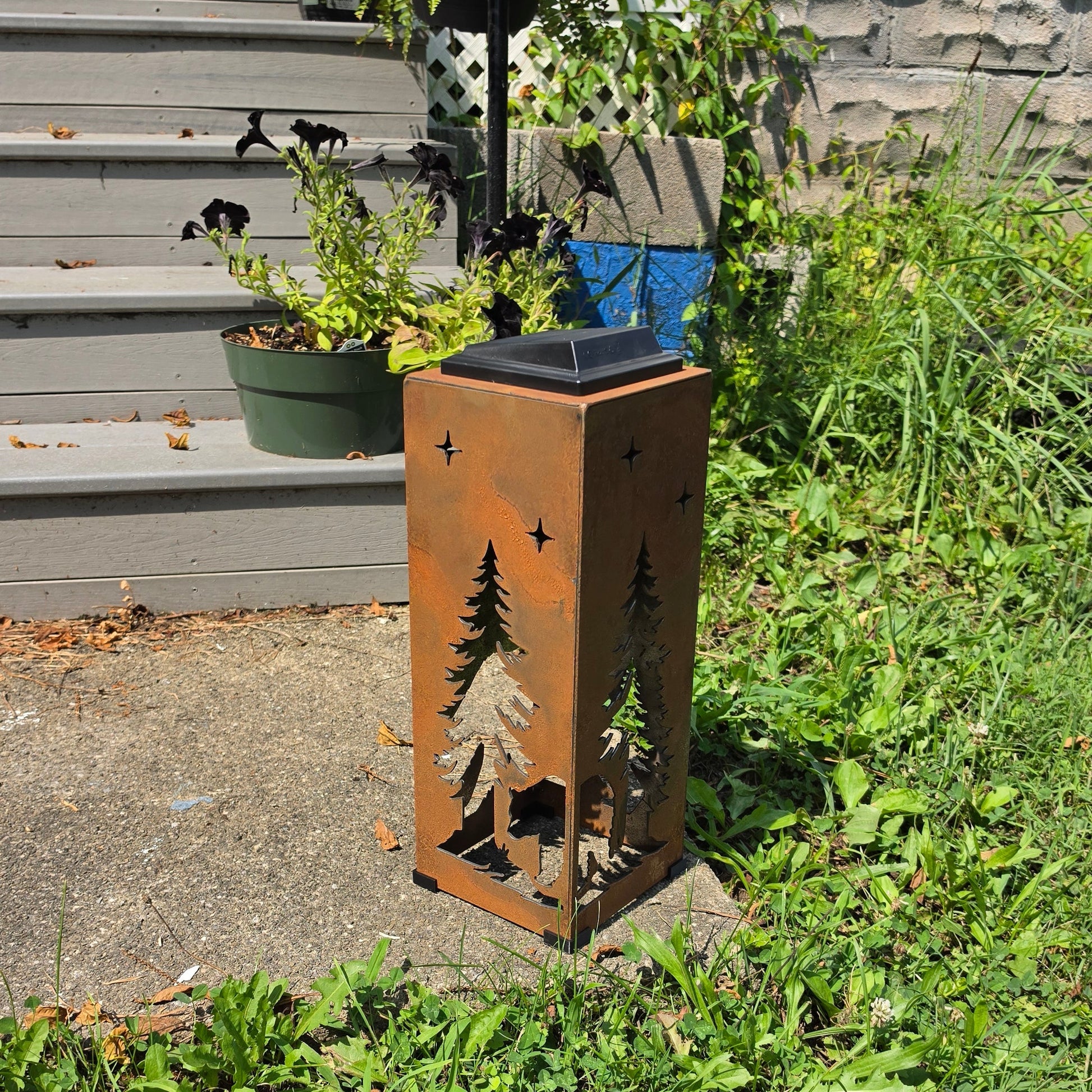 A rusted metal outdoor lantern with laser-cut tree and star designs, topped with a small solar panel, placed on a concrete path in a grassy yard near house steps.