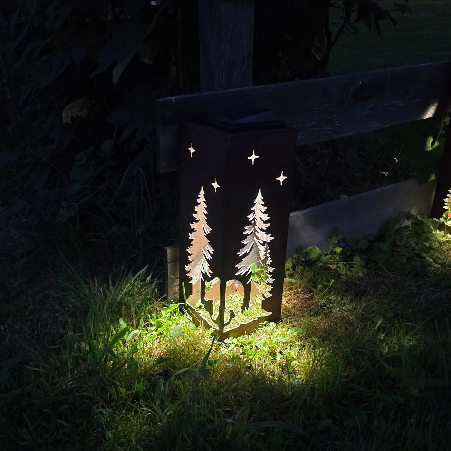 A dark outdoor ground lantern with laser-cut pine tree and star designs, illuminated at night on grass beside a wooden fence.