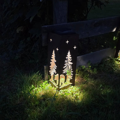 A dark outdoor ground lantern with laser-cut pine tree and star designs, illuminated at night on grass beside a wooden fence.