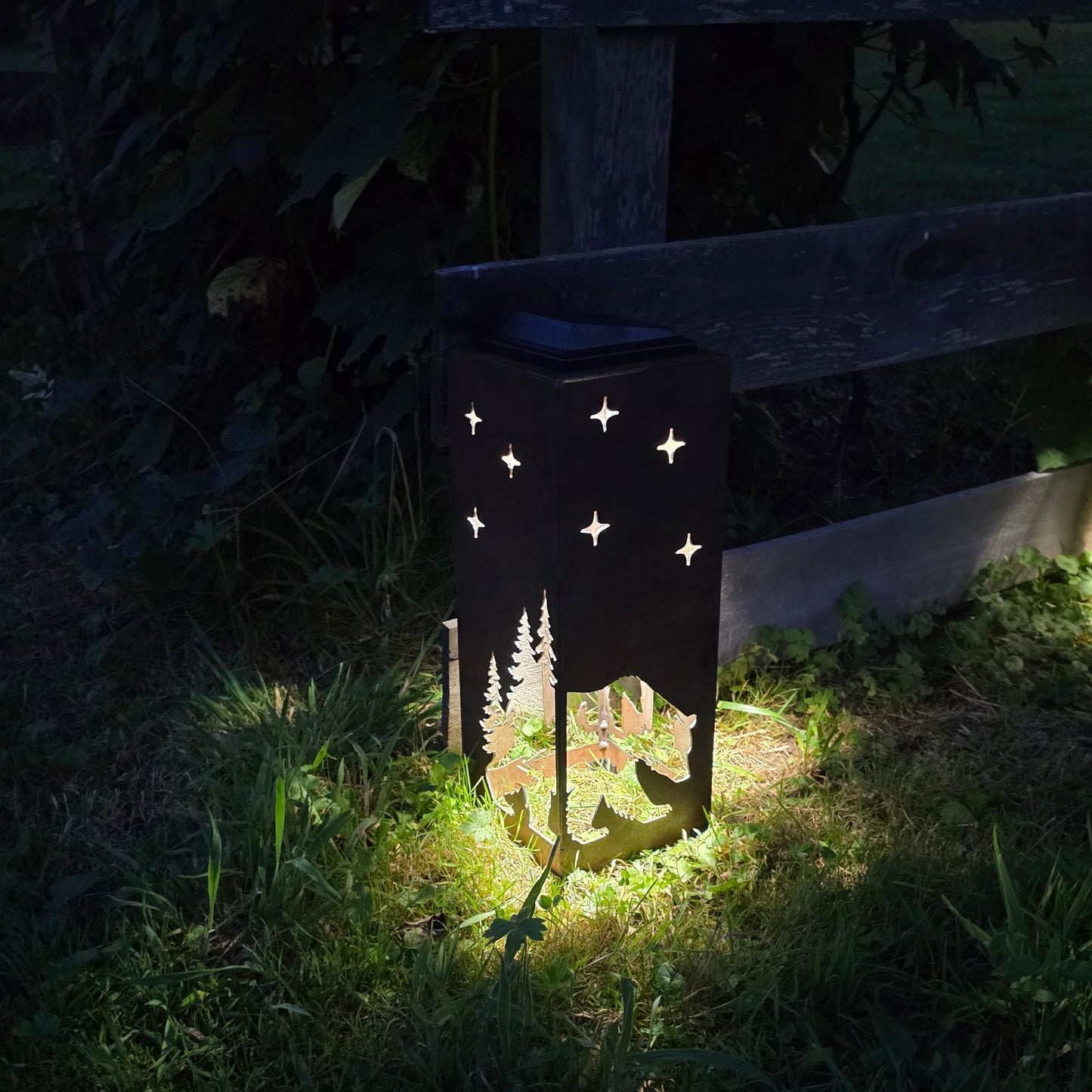 A rustic outdoor solar lantern with a forest silhouette and star cutout design, glowing with warm light on a grassy patch next to a wooden fence at night.