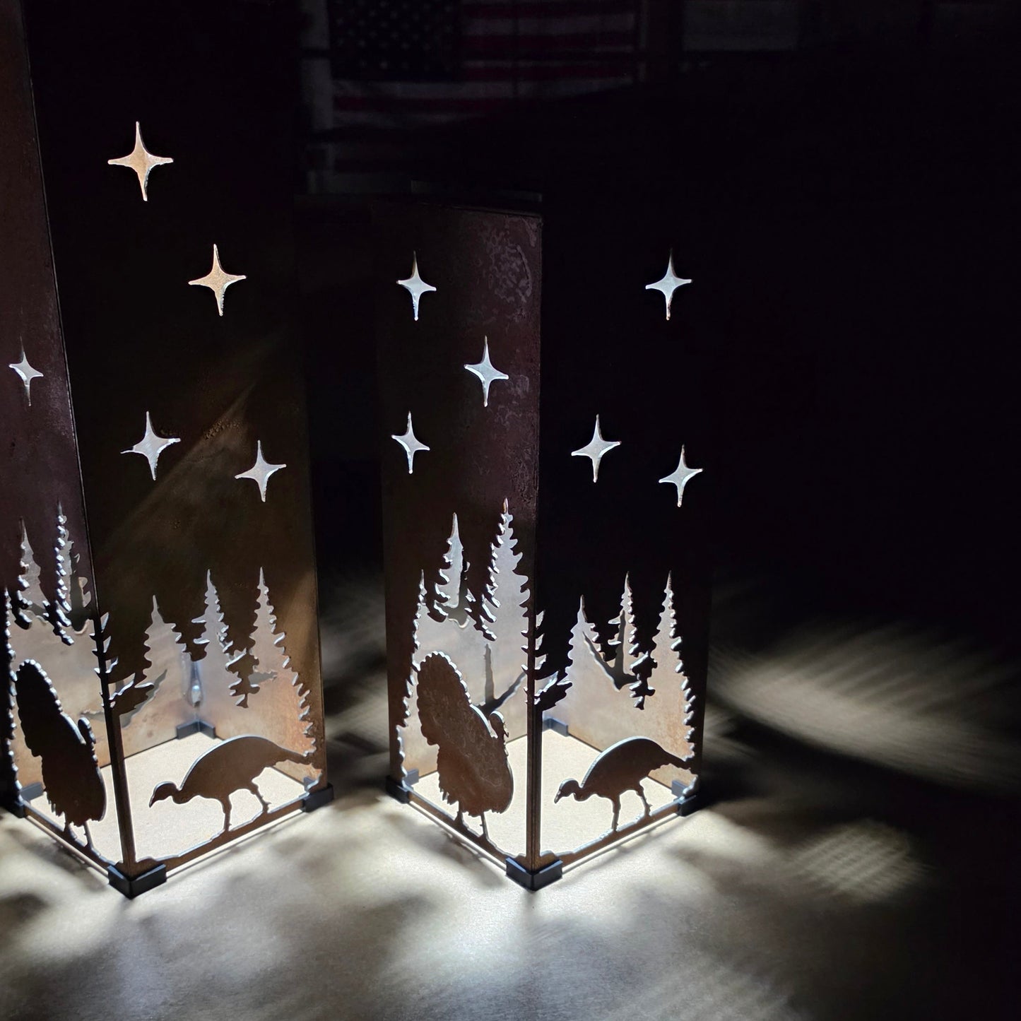 Two tall rectangular metal lanterns with laser-cut silhouettes of trees, turkeys, and stars, glowing with warm light in a dark room.