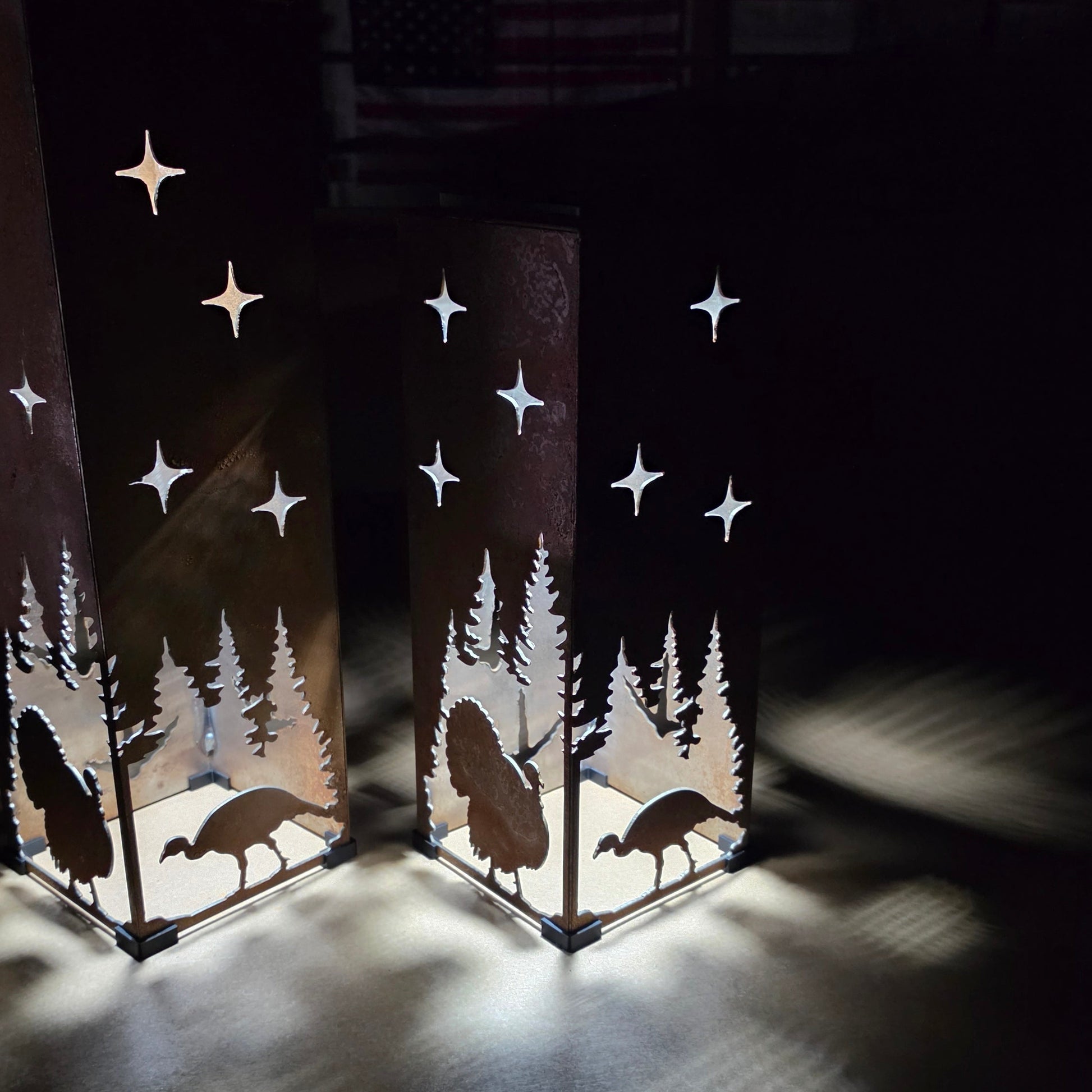 Two tall rectangular metal lanterns with laser-cut silhouettes of trees, turkeys, and stars, glowing with warm light in a dark room.