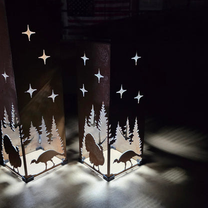 Two tall rectangular metal lanterns with laser-cut silhouettes of trees, turkeys, and stars, glowing with warm light in a dark room.