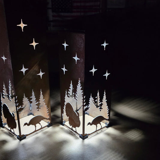 Two tall rectangular metal lanterns with laser-cut silhouettes of trees, turkeys, and stars, glowing with warm light in a dark room.