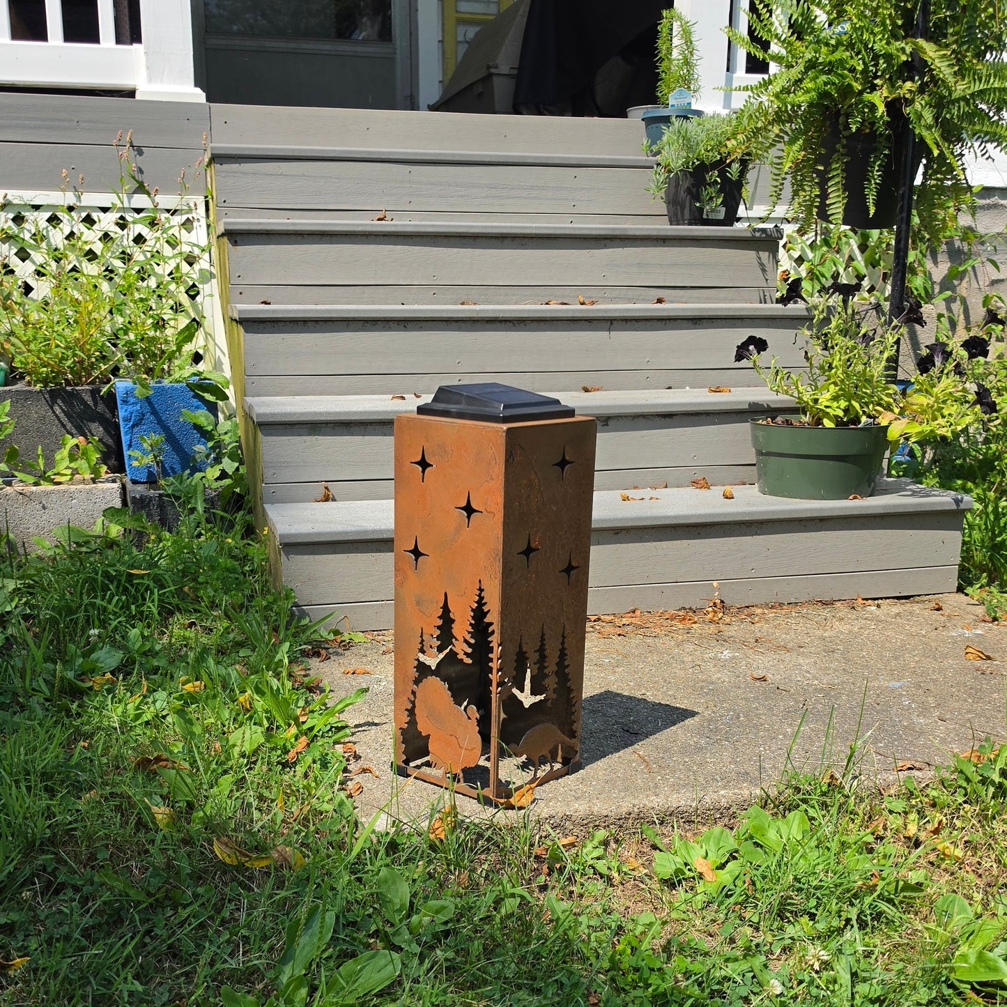 A rectangular metal outdoor lantern with a rusted copper-brown finish, featuring black silhouettes of pine trees and stars, placed on a concrete step in front of a house with gray steps and green grass.