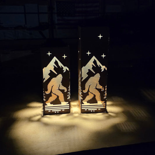 Two tall rectangular black lanterns with laser-cut silhouettes of a Bigfoot figure, mountains, and stars, glowing with warm light on a dark surface.