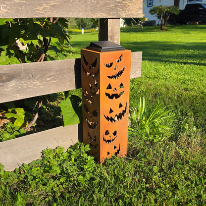A rust-orange metal Halloween yard light with laser-cut jack-o'-lantern faces, standing on grass next to a wooden fence in a backyard setting.
