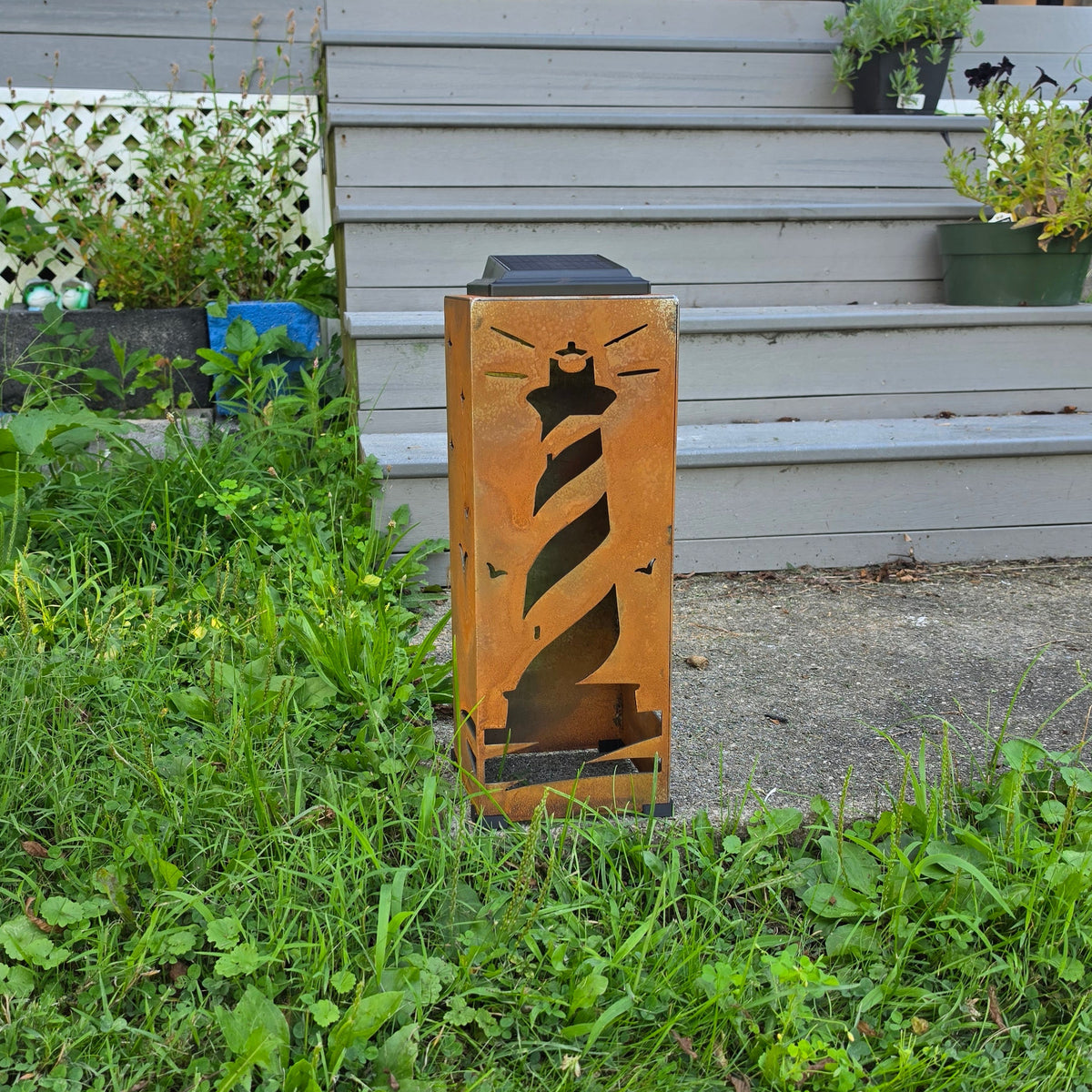 A rustic wooden outdoor light fixture shaped like a lighthouse, standing on a patch of green grass in front of a house porch.