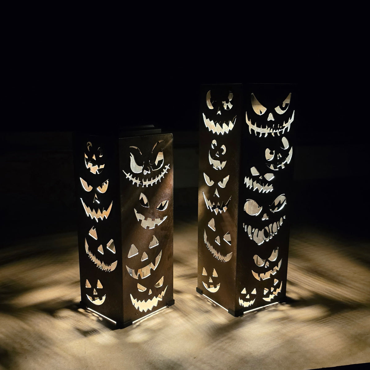 Two tall rectangular black lanterns with carved jack-o'-lantern cutouts, glowing from within on a dark surface against a black background.