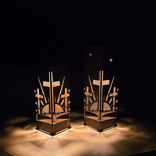 Two decorative metal candle holders with cutout religious symbols, including crosses, stars, and a book, shown lit on a dark surface against a black background.