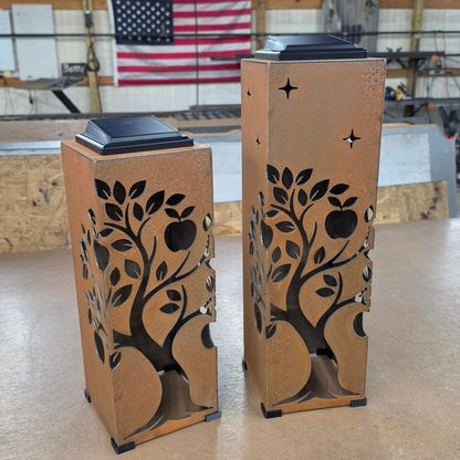 Two decorative metal lanterns with a tree-of-life cutout design and small solar panel tops, standing on a workshop floor in front of a blurred American flag and industrial ceiling.