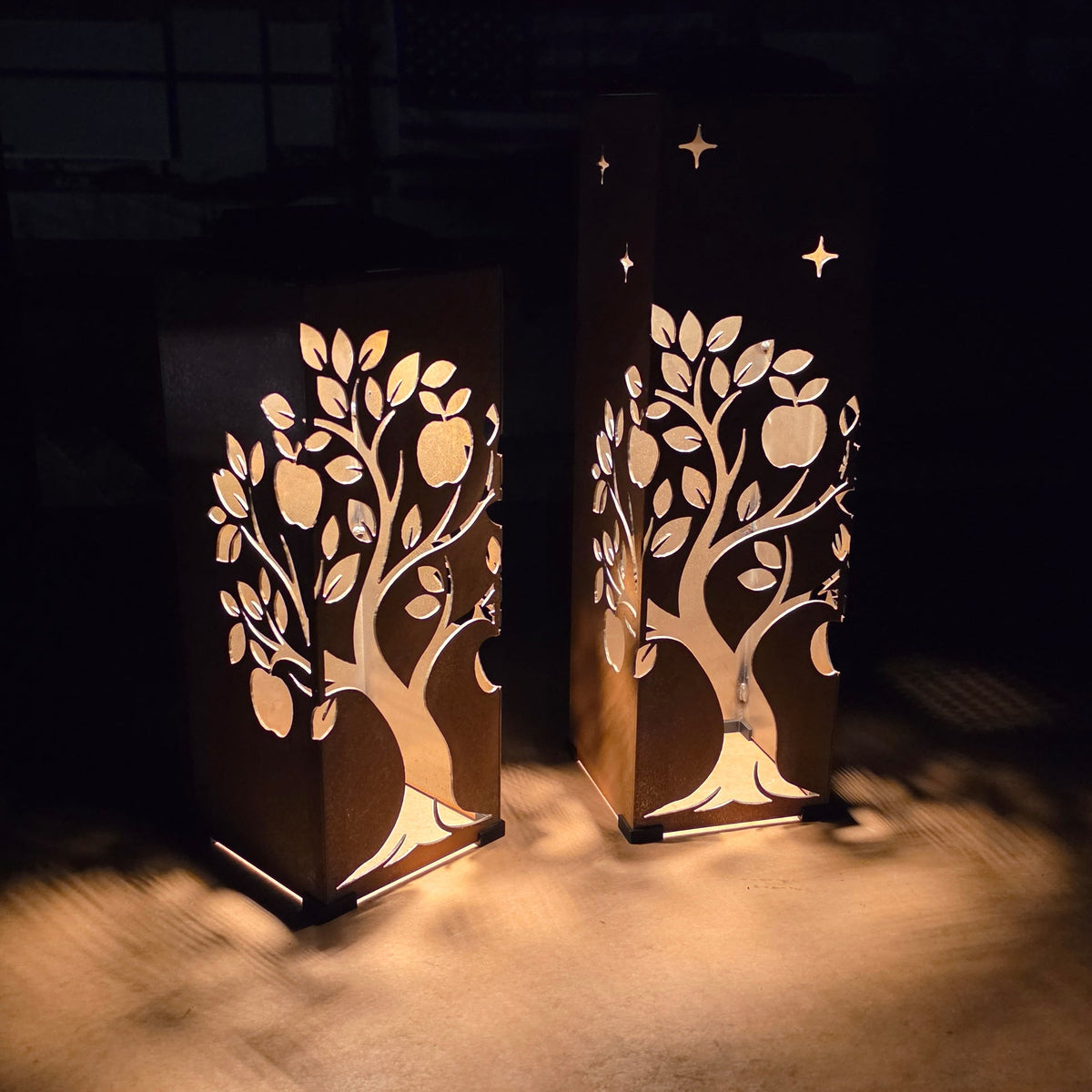 Two decorative metal lanterns with laser-cut tree of life and star patterns, glowing warmly on a dark surface.