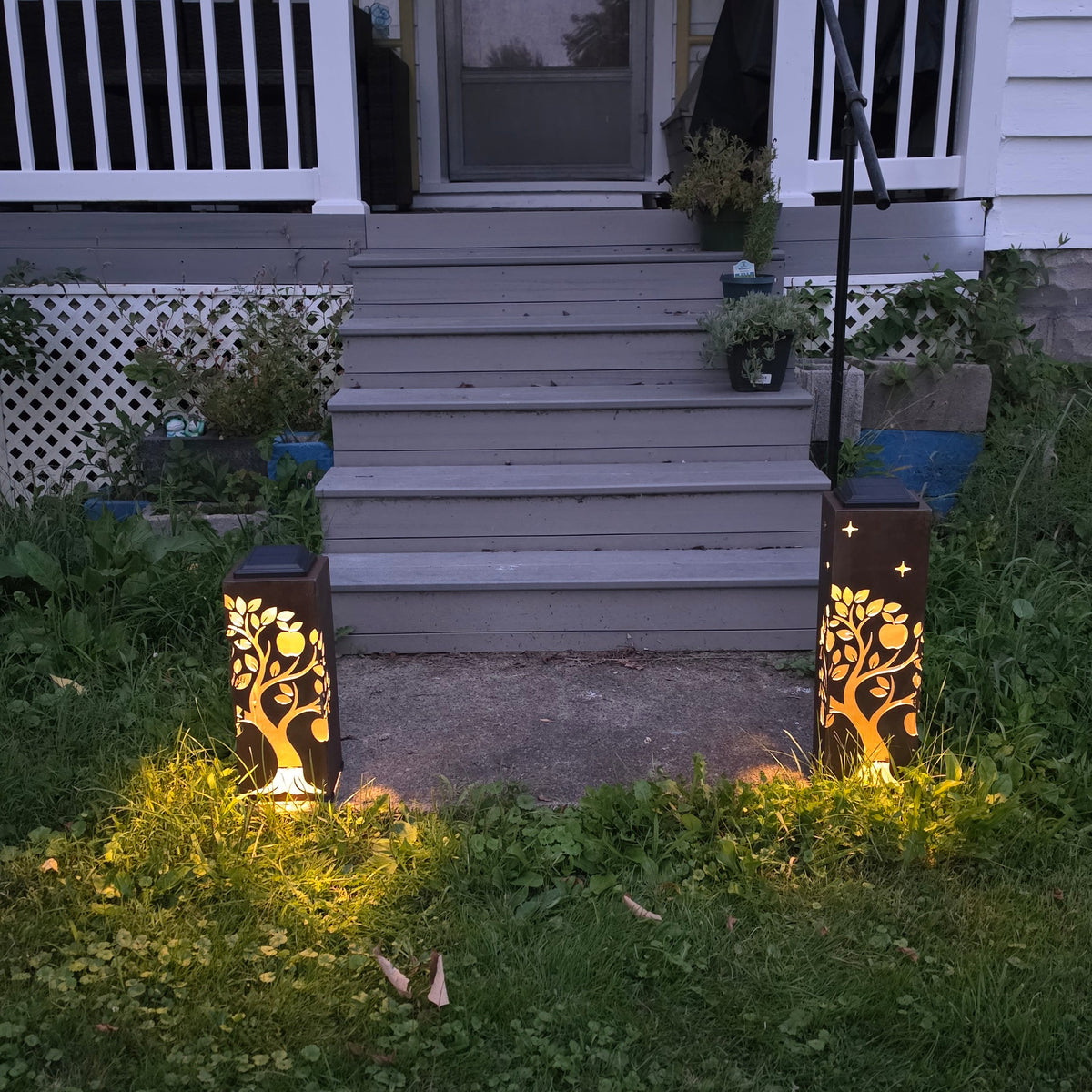 Two decorative outdoor solar lanterns with gold tree cutout designs are illuminated on either side of a front porch steps at dusk.