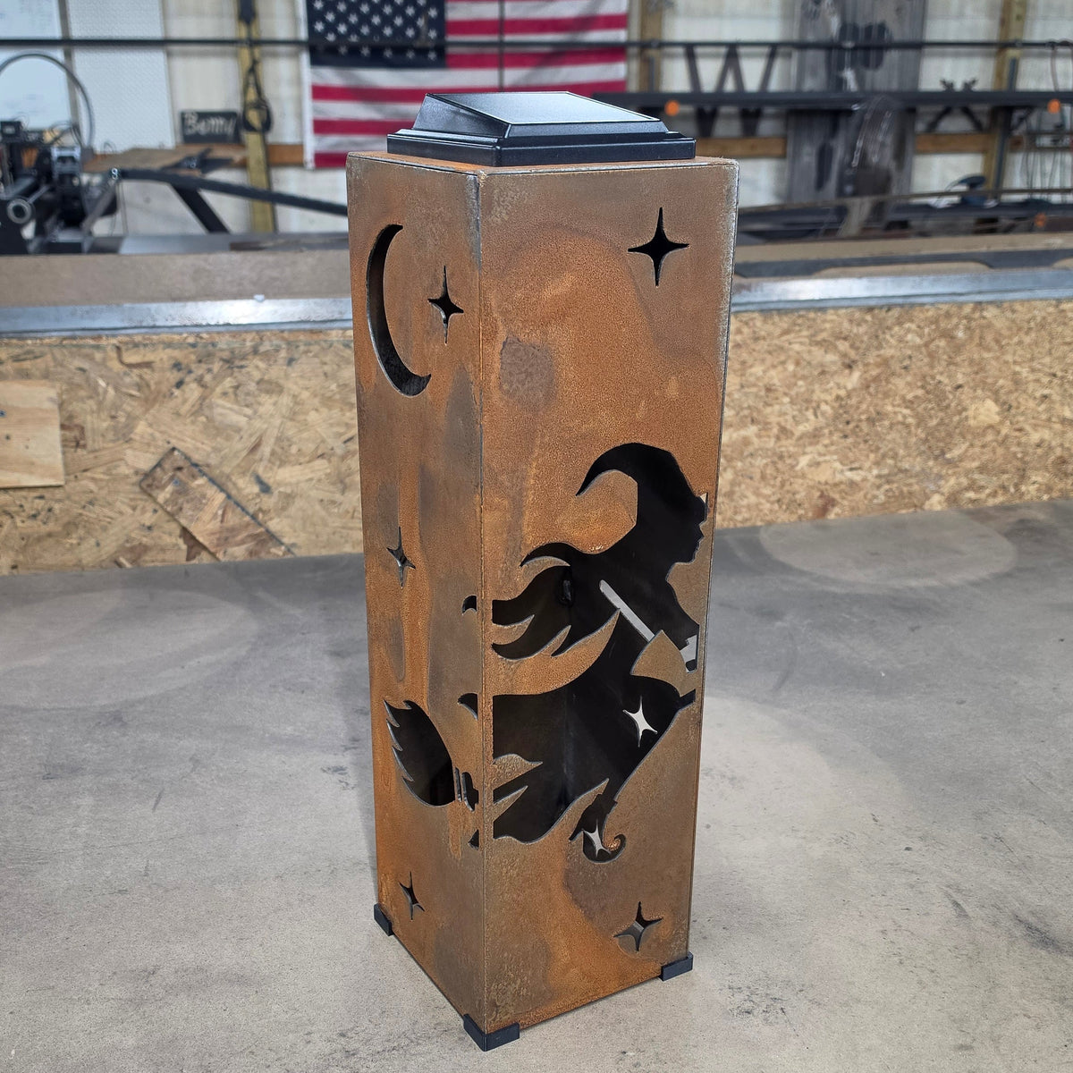 A rustic, weathered brown metal lantern with a witch silhouette, broom, stars, and crescent moon cutout, standing on a concrete floor in a workshop.