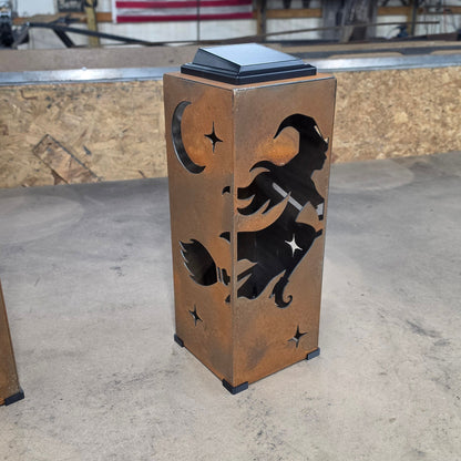 A rectangular metal sculpture or decorative container with a copper-toned finish, featuring black cutout designs of a witch riding a broomstick under a crescent moon and stars. It has a dark rectangular top and is photographed on a concrete floor inside a workshop with an American flag in the background.