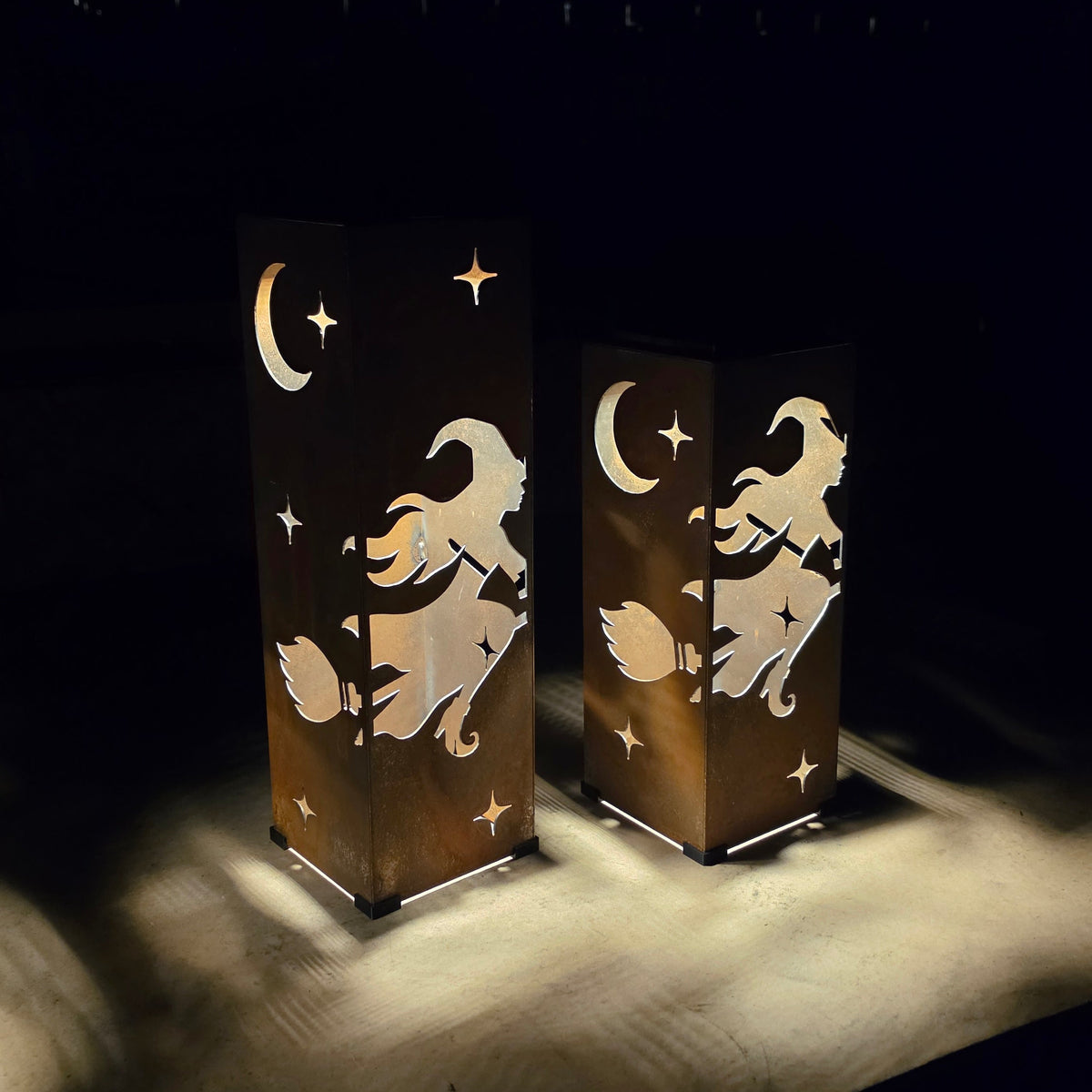Two rectangular decorative lanterns with a witch riding a broomstick and crescent moon design, casting warm light on a dark surface in a dim setting.