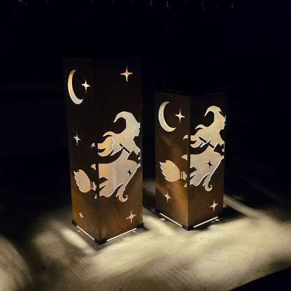 Two rectangular decorative lanterns with a witch riding a broomstick and crescent moon design, casting warm light on a dark surface in a dim setting.