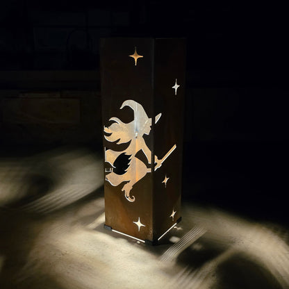 A rectangular wooden box or lamp with a cutout design of a witch riding a broomstick and small stars, shown glowing in a dark room and casting light onto the floor.
