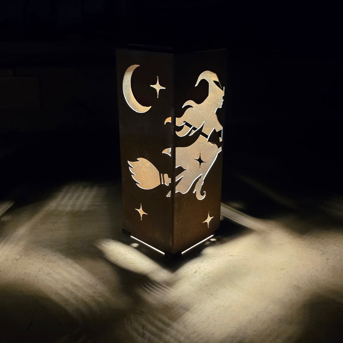 A dark rectangular decorative lamp or lantern with cutout designs of a witch riding a broomstick, a crescent moon, and stars, casting warm light onto a sandy surface in a dark setting.