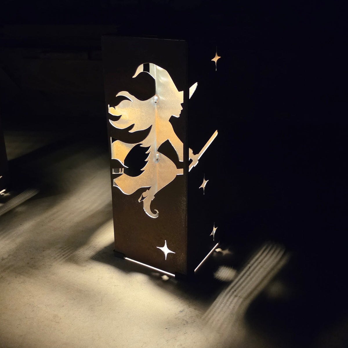 A dark metal outdoor light fixture featuring a laser-cut silhouette of a witch riding a broomstick with stars, casting a warm golden glow onto a dark patio floor.