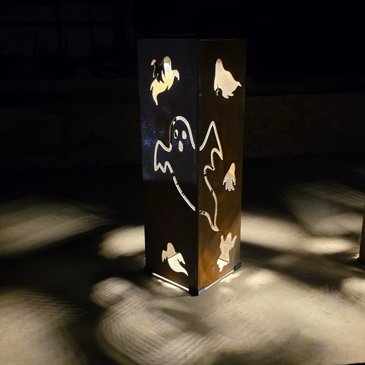 A tall rectangular metal lantern with ghost and bird cutouts, glowing warmly from within on a dark surface that casts soft shadows.