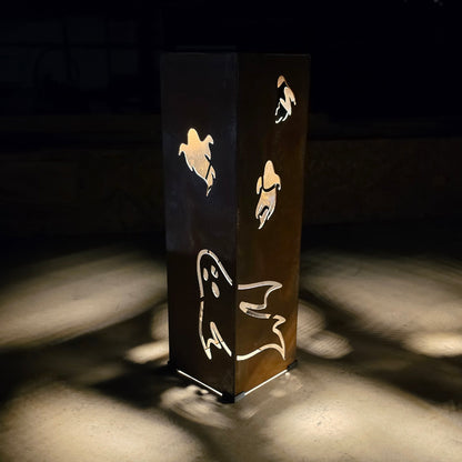A tall rectangular metal lantern with ghost cutouts, illuminated from within to cast soft light patterns on the surrounding dark ground.