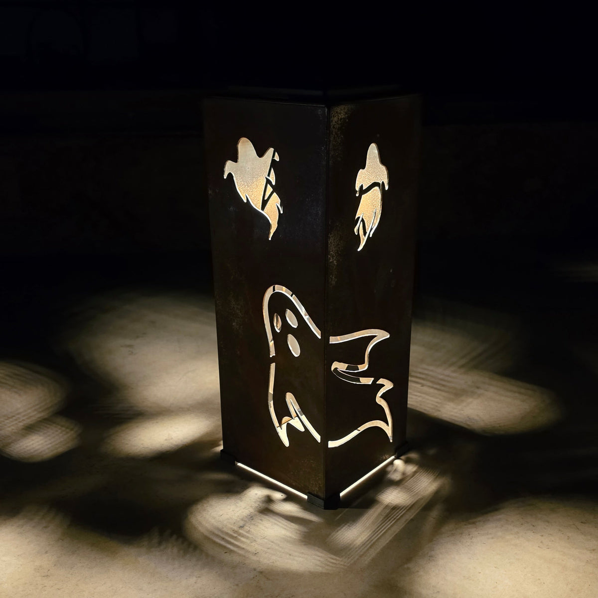 A dark rectangular lantern with ghost and bird cutout designs, casting warm light and shadows on a dark floor.