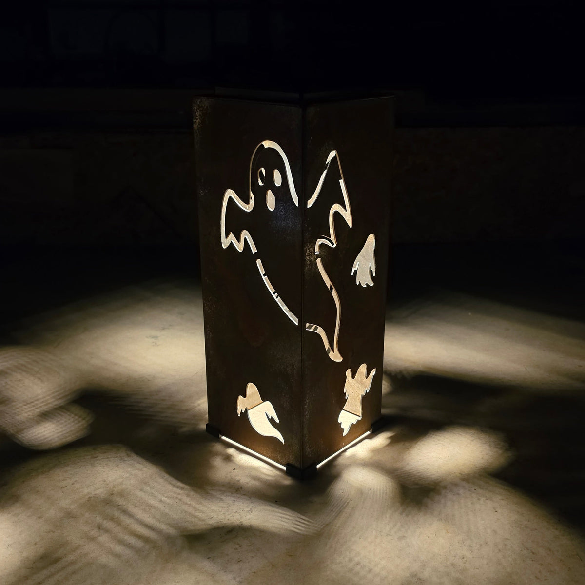 A dark metal lantern with ghost cutouts, glowing from within and casting dramatic shadows on a light-colored surface in a dark setting.