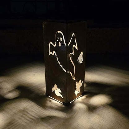A dark metal lantern with ghost cutouts, glowing from within and casting dramatic shadows on a light-colored surface in a dark setting.