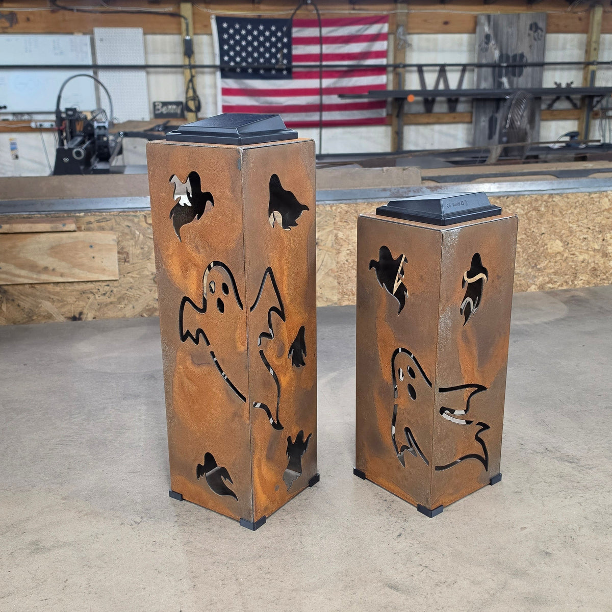 Two rustic metal lanterns with ghost cutouts and built-in LED lights, displayed on a workshop floor in front of a wooden wall with an American flag.