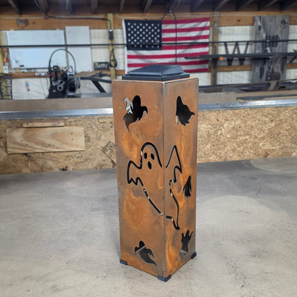 A rectangular metal lantern with a rusted orange-brown finish and black cutout designs of a ghost, bats, and birds, standing on a workbench in a workshop with an American flag in the background.