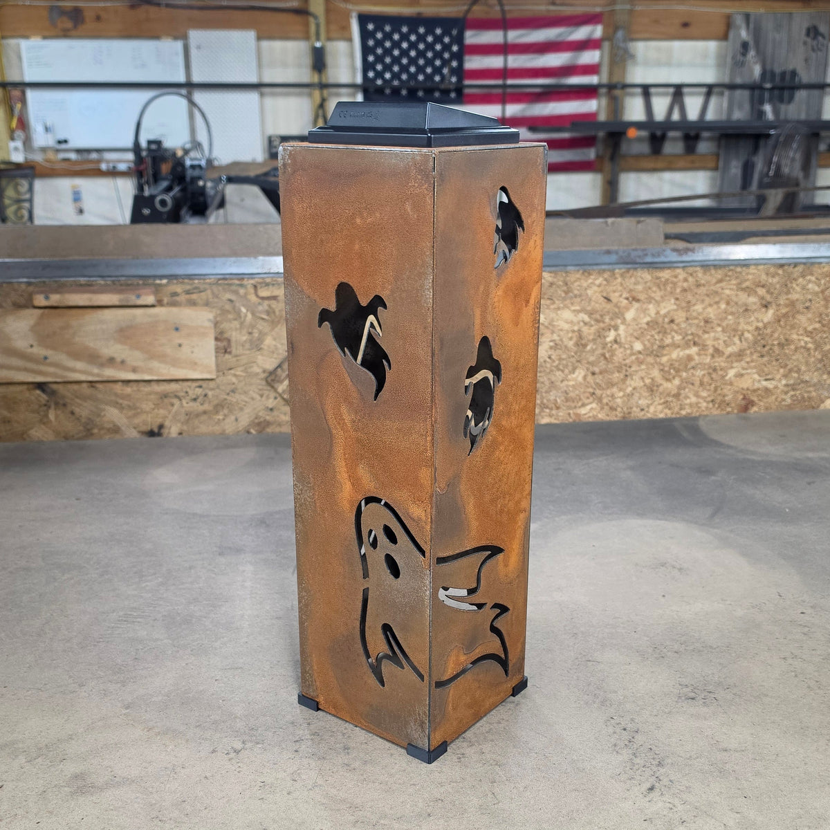 A rustic metal Halloween lantern with ghost and bird cutouts, standing on a workshop table with a garage-style background.