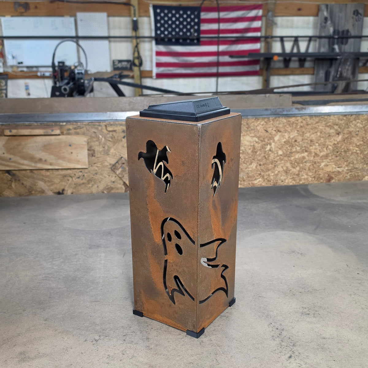 A rectangular metal container with a rusted copper-brown finish, featuring cutout ghost and bird designs on the front, topped with a black lid, set on a workshop table with an American flag in the background.
