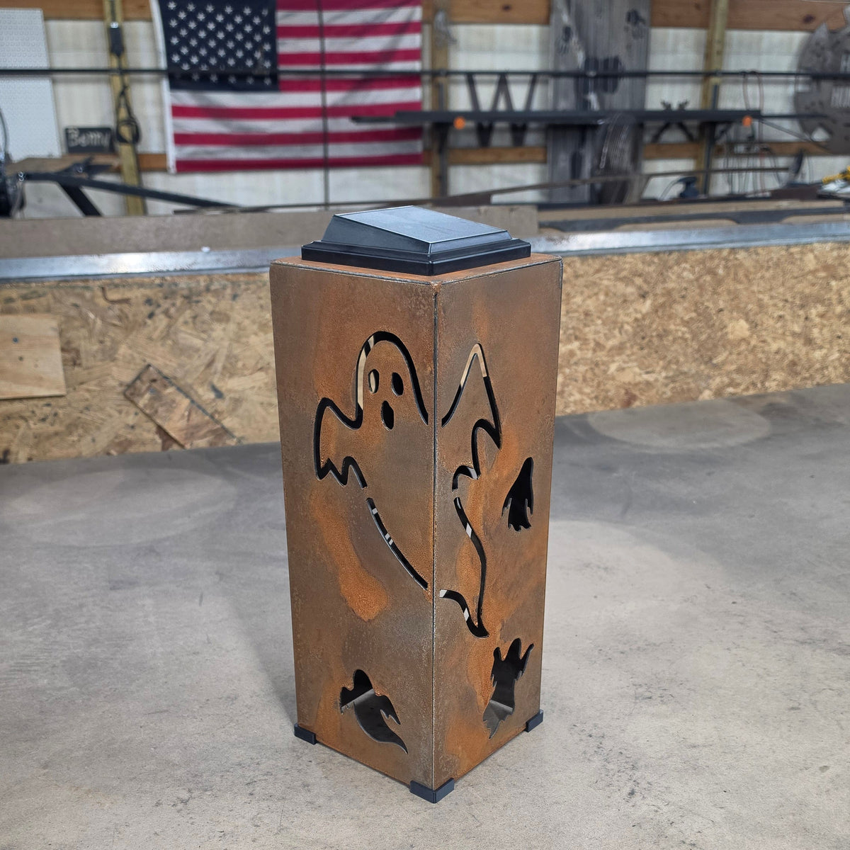 A rusted metal outdoor lantern with ghost cutouts, standing on a concrete workshop floor with an American flag and wooden beams in the background.