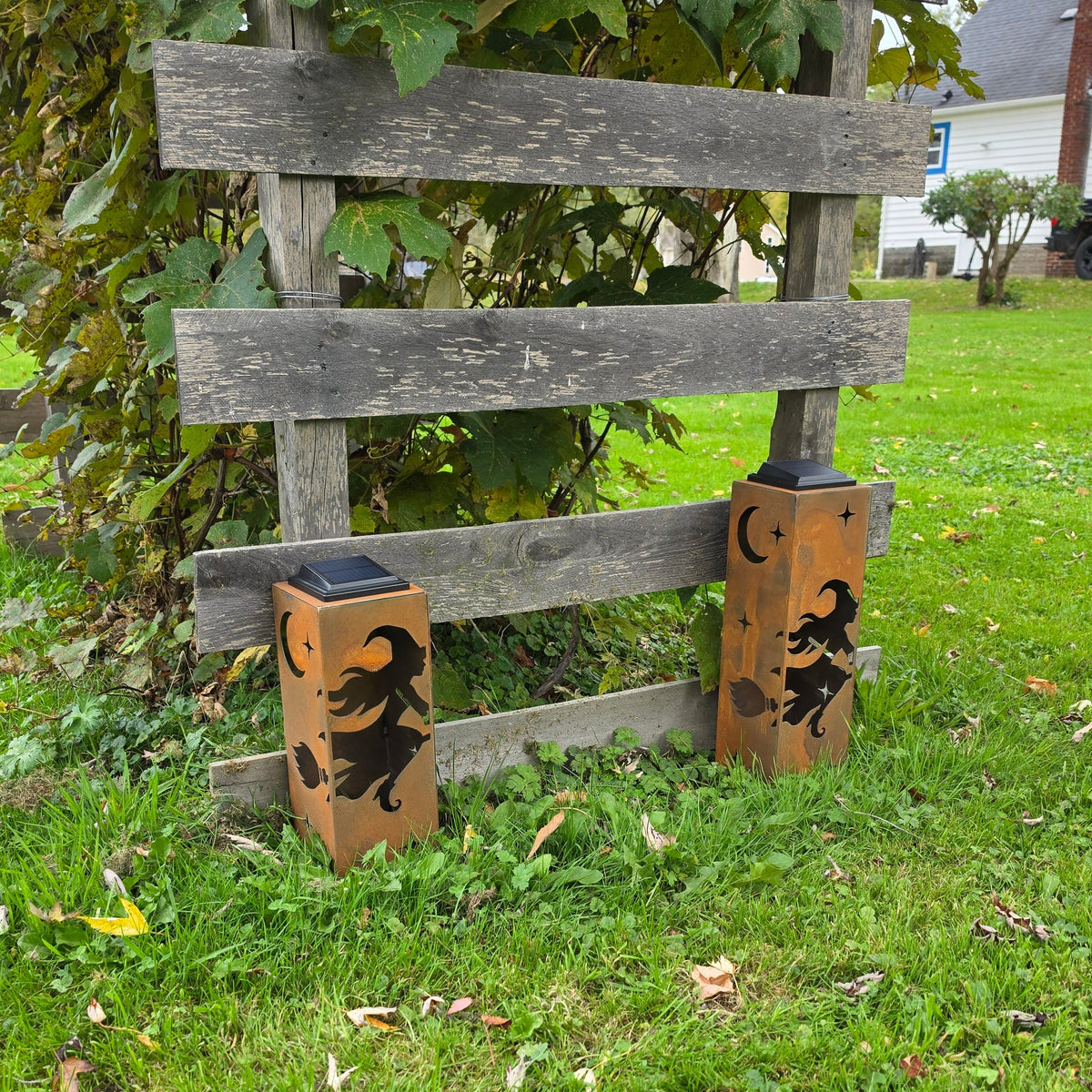 Two decorative outdoor lanterns with black witch and broomstick cutouts on a warm brown wood finish, placed on a green grassy lawn in front of a weathered wooden fence.