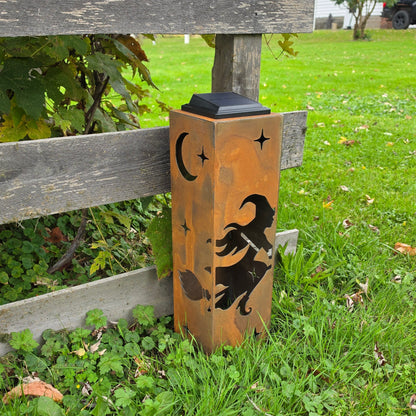 A rectangular outdoor solar light with a warm orange-brown finish, featuring black cutout designs of a witch riding a broomstick, a crescent moon, and stars. The light is shown standing on grass next to a wooden fence in a backyard setting.