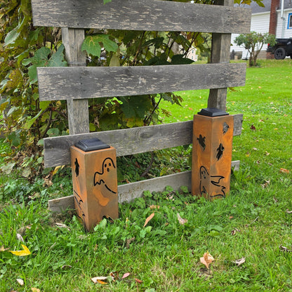 Two Halloween-themed metal ground lights with a rustic orange-brown finish, decorated with laser-cut ghost and spider designs, set on a green lawn next to a wooden fence.
