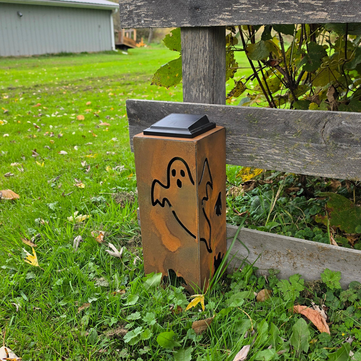 A rusted metal outdoor solar light shaped like a ghost, standing on grass next to a weathered wooden fence.