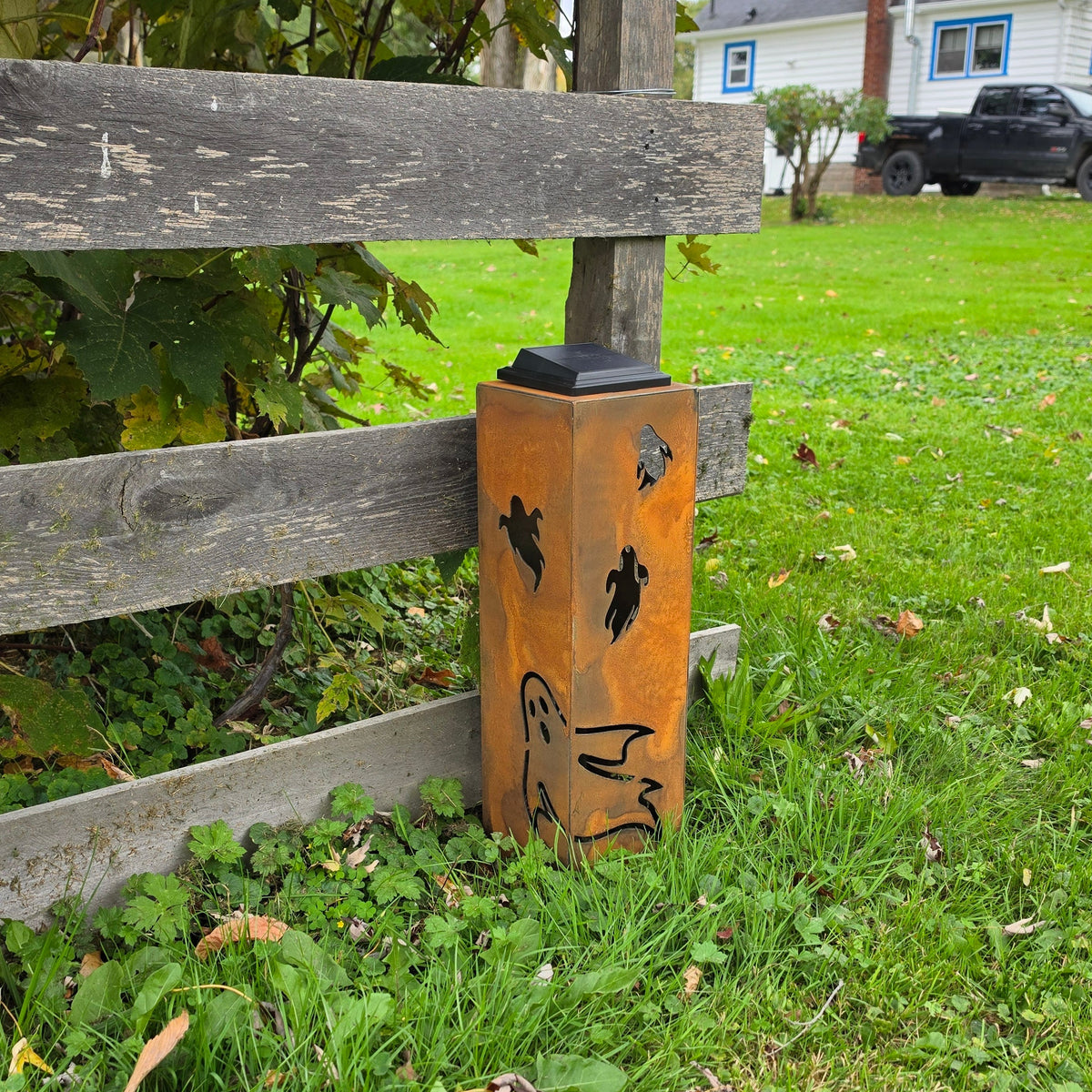 A rust-orange metal fence post cap with black decorative cutouts, mounted on a wooden fence post in a grassy yard.