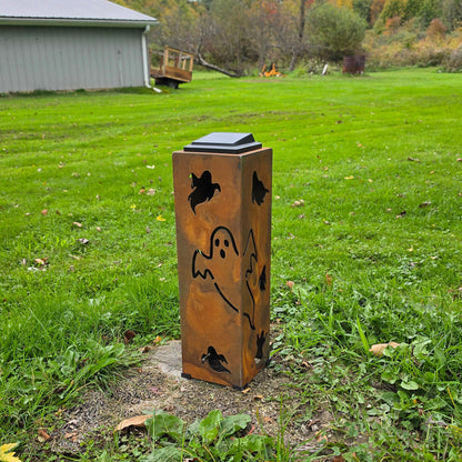 A rectangular metal outdoor light post with a rusted copper finish, decorated with black silhouettes of a ghost, bats, and a spider, standing on a patch of dirt in a grassy yard.