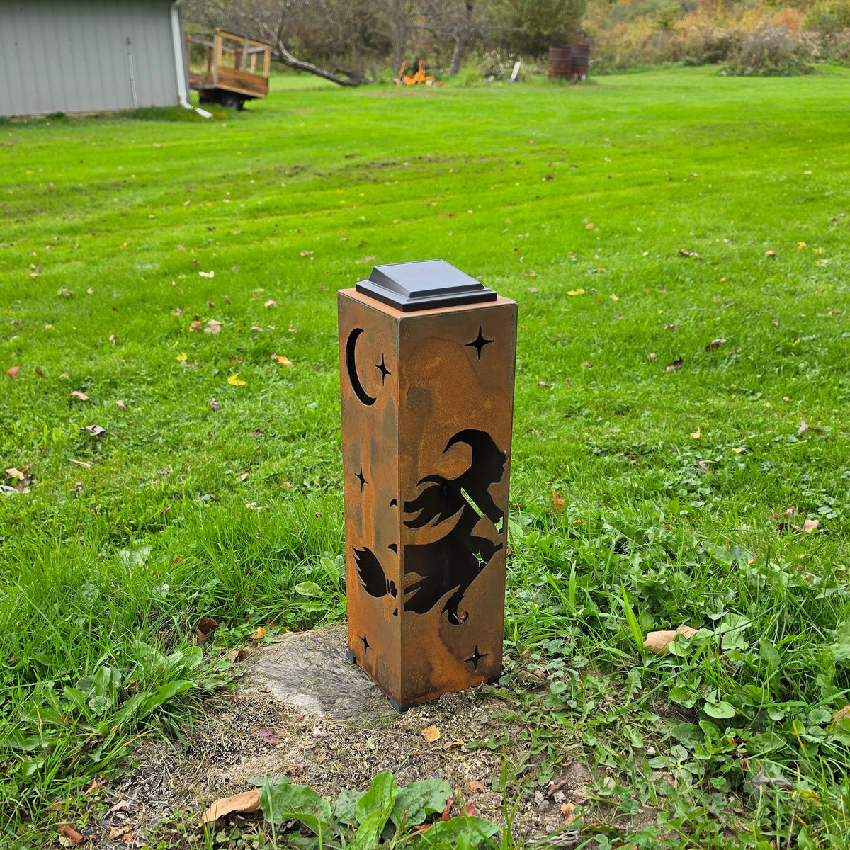 A rustic wooden outdoor light post featuring a witch silhouette and moon cutout, set on a grassy lawn with a small solar panel on top.