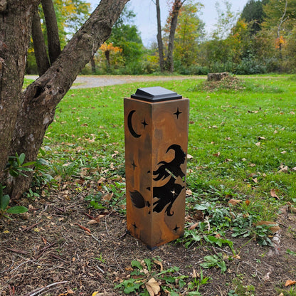 A rectangular outdoor light post with a weathered copper-brown finish, featuring laser-cut black silhouettes of a witch riding a broomstick under a crescent moon and stars. The post is situated outdoors on a grassy lawn near a tree trunk.