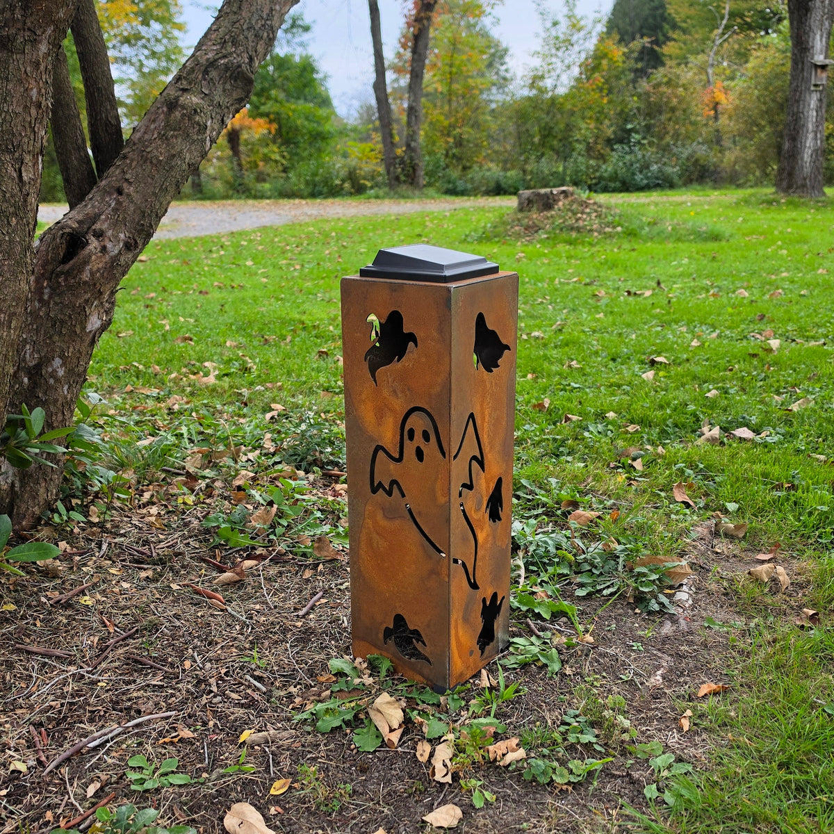 A rusted metal outdoor trash can with a black lid, decorated with cutout designs of a ghost, mountain, and butterflies, situated on a grassy park path near trees.