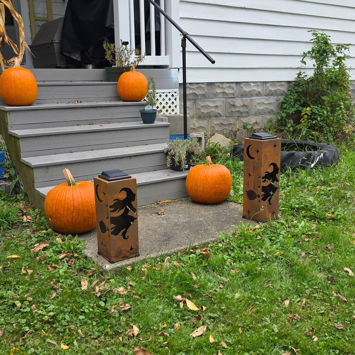 Outdoor front yard of a house with gray steps decorated with orange pumpkins and two wooden witch-themed bollards on the lawn.