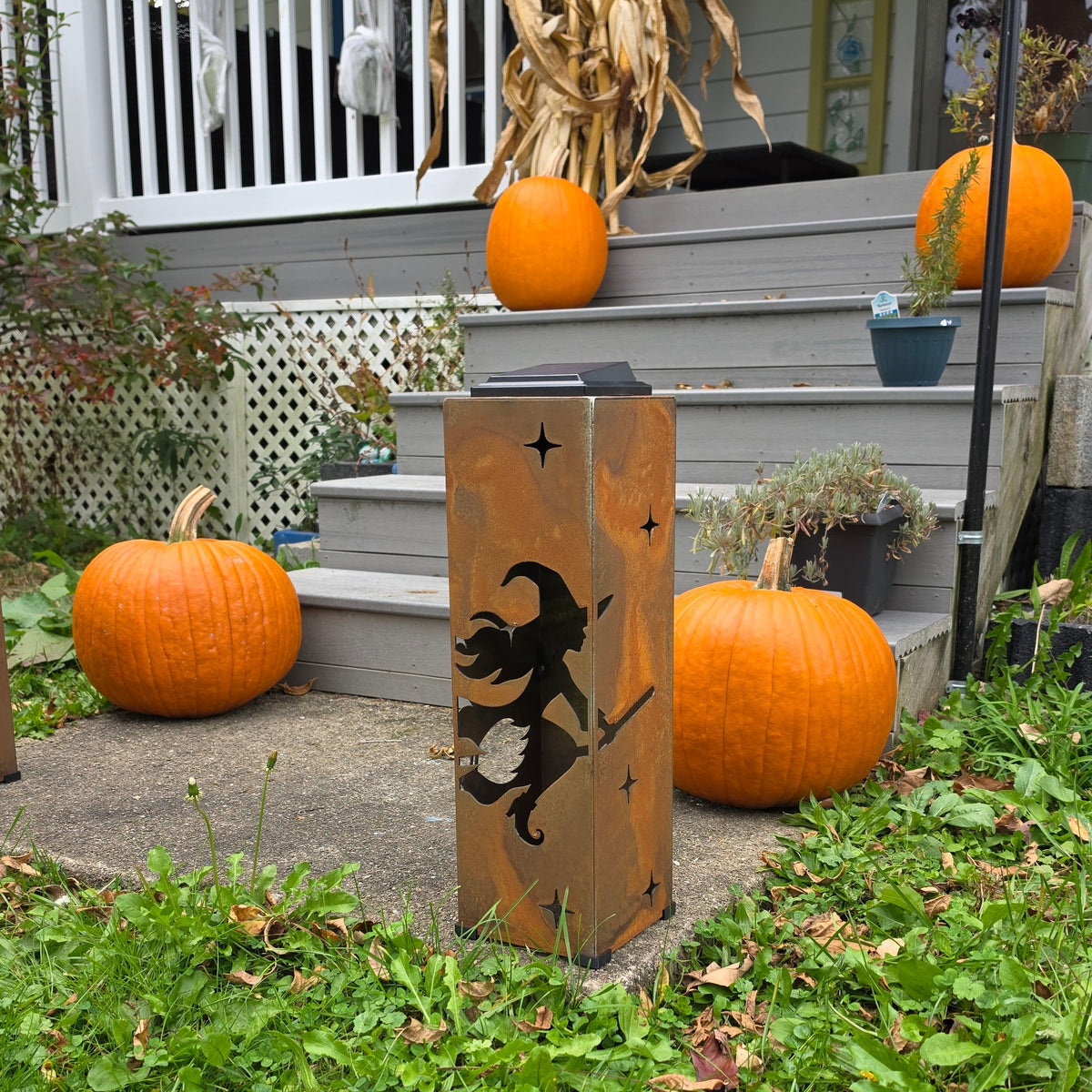 A tall rectangular wooden Halloween light with a black silhouette of a witch riding a broomstick, placed on a front porch steps surrounded by pumpkins and grass.