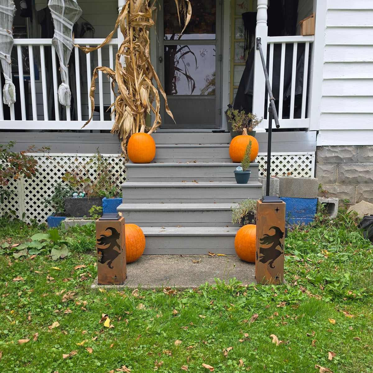 Outdoor front porch and lawn decorated for Halloween, with orange pumpkins on the steps and porch, a corn stalk, spider web decorations, and two wooden gate signs with a witch silhouette on a grassy lawn.