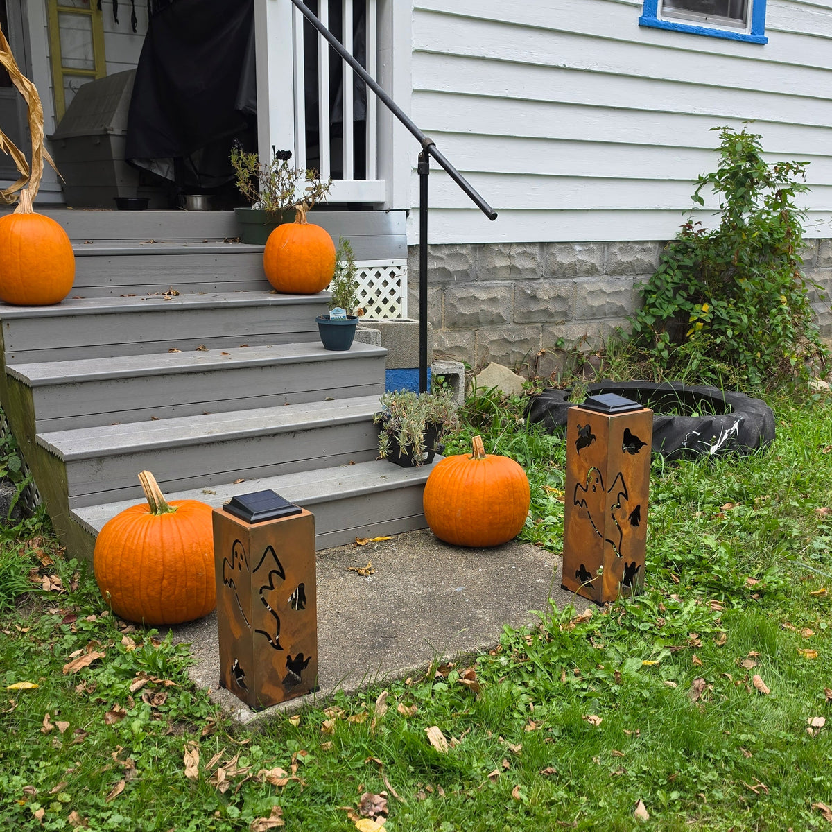 Outdoor front yard scene with a house porch, gray steps, several orange pumpkins, and two decorative lanterns placed on the grass and steps.