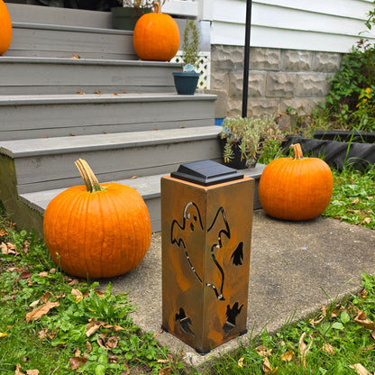 A rectangular outdoor solar light with a rusted copper finish and black bat silhouette cutouts, placed on a concrete path in a grassy yard with pumpkins nearby.
