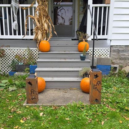 Outdoor front porch decorated for Halloween with orange pumpkins on the steps, a dried corn stalk, spider webbing, and two wooden gate posts with carved bats and the word 'Halloween' on a grassy lawn.